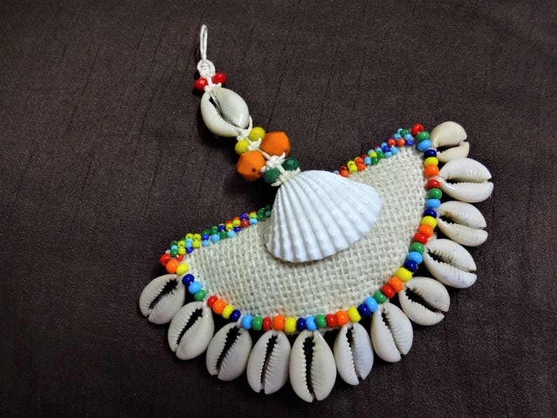 May include: A white seashell with a burlap background, surrounded by a colorful beaded trim and smaller white seashells.