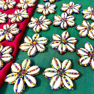 Puede incluir: Una colección de adornos en forma de flor hechos con conchas de cauri blancas y cuentas de semillas vibrantes y multicolores. Cada pieza presenta seis conchas dispuestas en un patrón circular, con cuentas que bordean los pétalos y el centro.