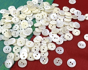 May include: A collection of small, round, off-white buttons with two holes each. The buttons are scattered on a red and green fabric background, creating a visually appealing arrangement. These buttons are ideal for sewing and craft projects.