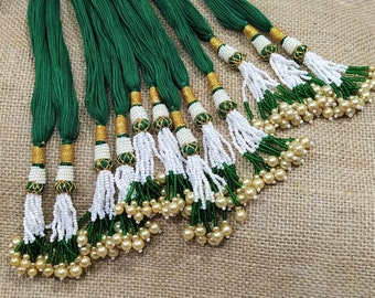 May include: A group of green and white beaded tassels with gold accents. The tassels are arranged in a row on a burlap background.