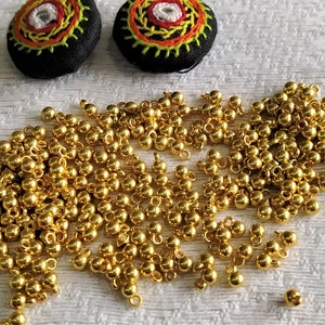 May include: A large quantity of gold-coloured metal beads with a loop on each bead. The beads are scattered on a white surface.