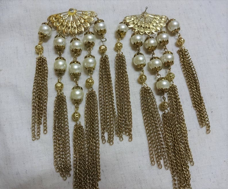 K&ouml;nnte beinhalten: Zwei goldfarbene Haarklammern mit Perlen und herabh&auml;ngenden goldenen Ketten. Die Klammern haben ein f&auml;cherf&ouml;rmiges Design.