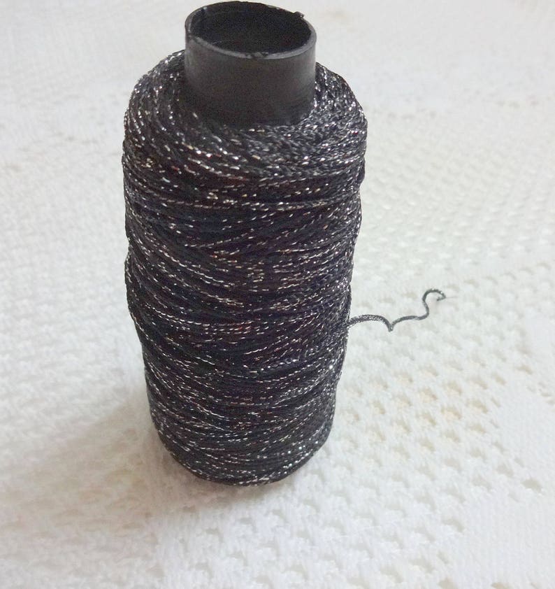 Puede incluir: Una bobina de hilo negro con detalles met&aacute;licos plateados. El hilo est&aacute; enrollado alrededor de una bobina de pl&aacute;stico negro.