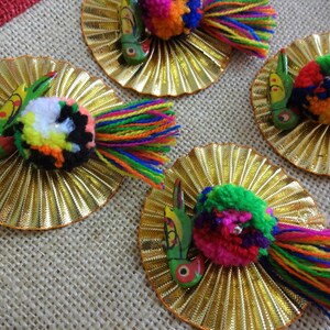 May include: Four colorful, handmade, wooden bird ornaments with pom-poms and fringe. Each ornament is attached to a gold, circular, decorative base.