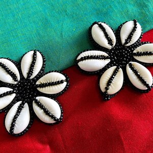 May include: Two flower-shaped accessories, each composed of six white seashells with black beaded centers and edges. The accessories are set against a red and green background, creating a striking contrast.