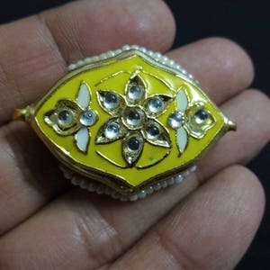 May include: A yellow and gold decorative jewelry piece with a floral design. The piece has a yellow enamel base, gold trim, and is adorned with clear gemstones and small white pearls. The design is symmetrical and ornate.