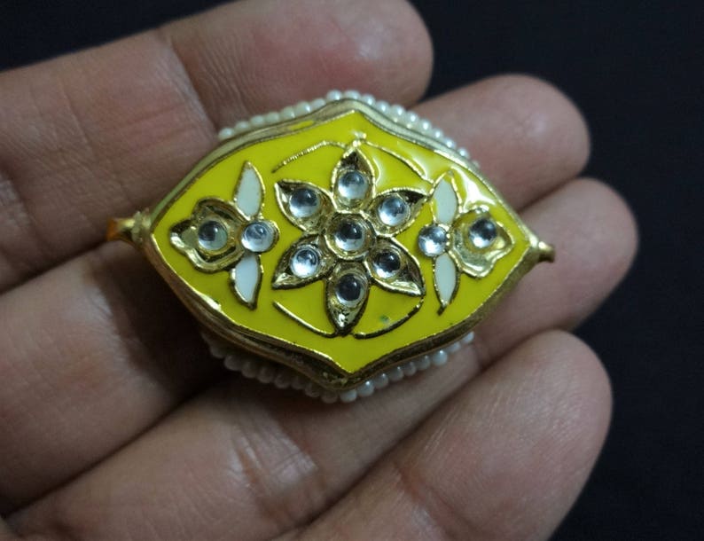 May include: A yellow and gold decorative jewelry piece with a floral design. The piece has a yellow enamel base, gold trim, and is adorned with clear gemstones and small white pearls. The design is symmetrical and ornate.