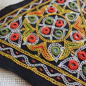 May include: A black fabric triangle with intricate embroidery in white, yellow, red, and green thread. The embroidery features a repeating pattern of circles and leaves.