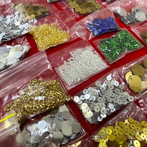 May include: Assortment of sequins and beads in various colors and shapes, packaged in clear plastic bags. The collection includes silver, gold, and iridescent sequins, along with gold, silver, and green beads. Ideal for crafting and embellishment projects.