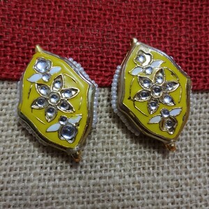 May include: A pair of yellow and gold earrings with a floral design. Each earring features a central flower with clear, sparkling accents and white petal details. The edges are lined with small, white beads. The earrings are set against a textured, neutral background.