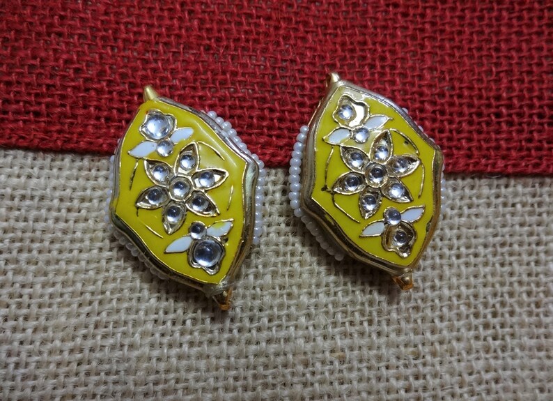 May include: A pair of yellow and gold earrings with a floral design. Each earring features a central flower with clear, sparkling accents and white petal details. The edges are lined with small, white beads. The earrings are set against a textured, neutral background.