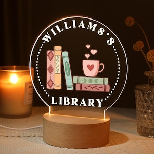 Puede incluir: Una luz nocturna de acrílico blanco con una base de madera. La luz presenta un diseño de libros apilados, una taza de café y el texto "William's Library".