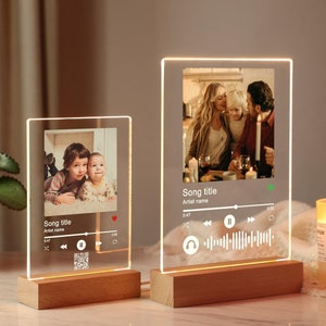 Op de afbeelding: Twee verlichte fotolijsten met een houten basis. De lijsten tonen foto's van families en hebben een muziekspelerinterface met een songtitel, artiestennaam en een voortgangsbalk. De lijsten zijn verlicht met warm wit licht.