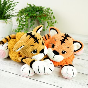 May include: Two crocheted tiger plushies, one orange and one yellow, with black stripes and white paws. The tigers are lying down with their heads together.