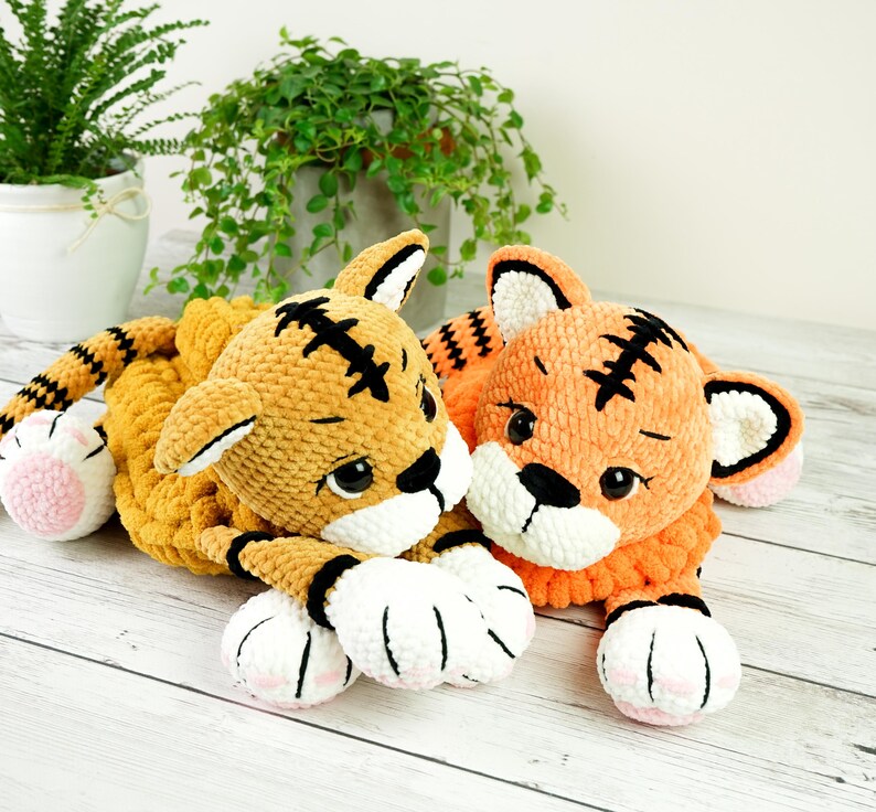 May include: Two crocheted tiger plushies, one orange and one yellow, with black stripes and white paws. The tigers are lying down with their heads together.