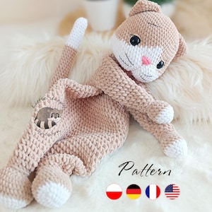 May include: A handmade crochet cat doll in soft beige with white accents. The cat has a white face, pink nose, and black eyes. The tail and paws are white. The word "Pattern" is written on the bottom right. The doll is lying on a white fluffy surface.