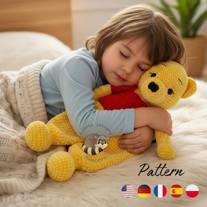 May include: A crocheted yellow bear with a red shirt is being hugged by a child. The bear has black eyes and a black nose. The bear is lying on a white fluffy surface. The word "Pattern" is written in black text.
