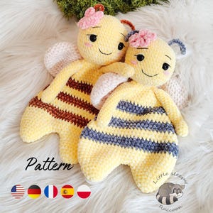 Puede incluir: Dos peluches de abeja hechos a mano con ganchillo. Las abejas son amarillas con rayas marrones y azules, detalles de flores rosas y caras sonrientes. La palabra "Pattern" está escrita en la parte inferior izquierda. Los peluches están sobre una superficie blanca y esponjosa.