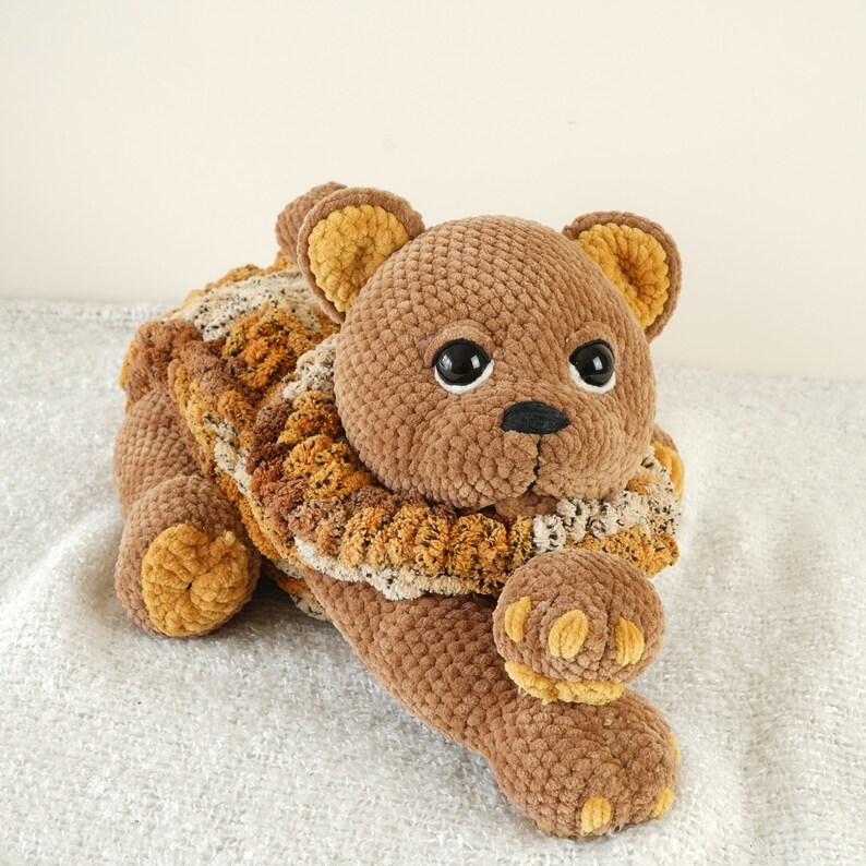 May include: A brown crocheted teddy bear with a brown and yellow crocheted collar. The bear has black eyes and a black nose.