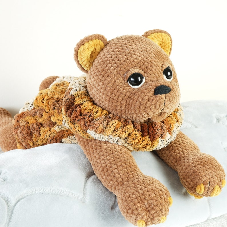 May include: A brown crocheted lion plush toy with a yellow mane and black eyes. The lion is lying on a white fluffy surface.