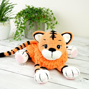 May include: A crocheted orange and black tiger plush toy with white and pink accents. The toy is lying on a white wooden surface.
