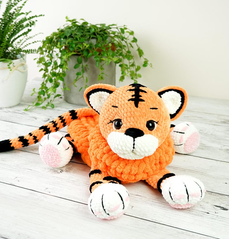 May include: A crocheted orange and black tiger plush toy with white and pink accents. The toy is lying on a white wooden surface.
