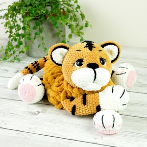 May include: A crocheted tiger plush toy with orange and black stripes, white paws, and pink paw pads. The toy is lying on a white wooden surface.