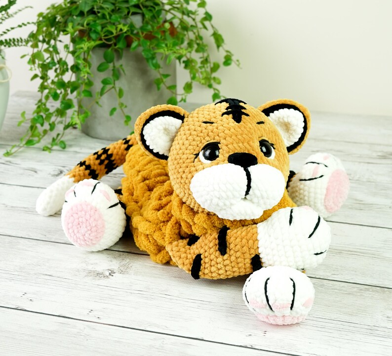 May include: A crocheted tiger plush toy with orange and black stripes, white paws, and pink paw pads. The toy is lying on a white wooden surface.