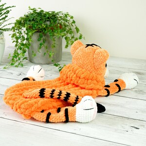 May include: A crocheted tiger plush toy with orange and black stripes. The toy has a white face with black eyes and a pink nose. The toy is lying on a white wooden surface.