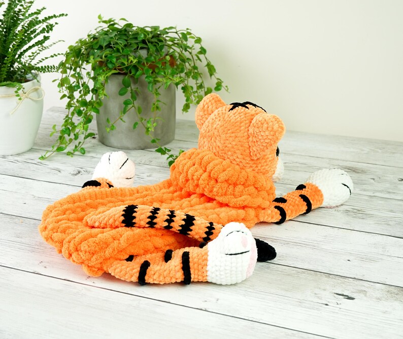 May include: A crocheted tiger plush toy with orange and black stripes. The toy has a white face with black eyes and a pink nose. The toy is lying on a white wooden surface.
