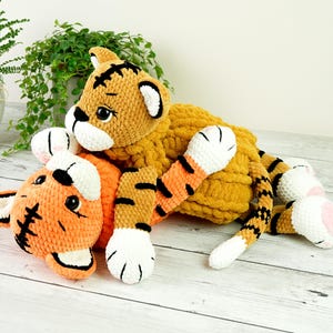May include: Two crocheted tiger plushies, one orange and one yellow, with black stripes and white accents. The orange tiger is lying on its back with its paws in the air, while the yellow tiger is lying on top of it.