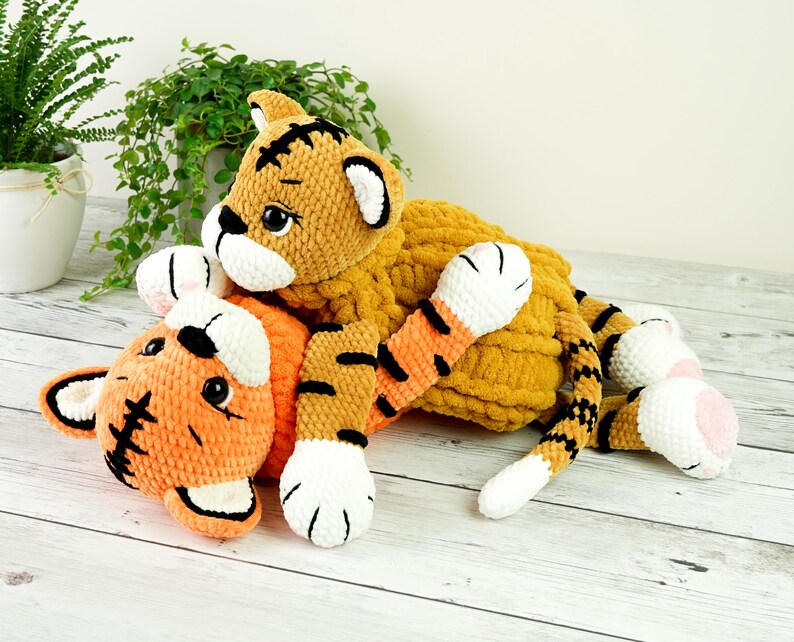 May include: Two crocheted tiger plushies, one orange and one yellow, with black stripes and white accents. The orange tiger is lying on its back with its paws in the air, while the yellow tiger is lying on top of it.