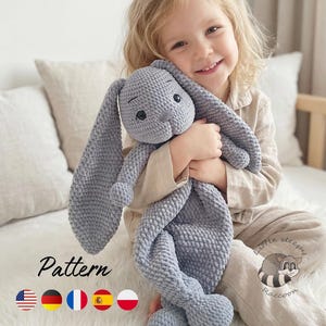 May include: A gray crocheted plush toy with long floppy ears, embroidered eyes, and a textured yarn surface. The toy features a soft, stuffed head and a flat, blanket-like body. The image includes the text Pattern and a Little Sleepy Raccoon logo.