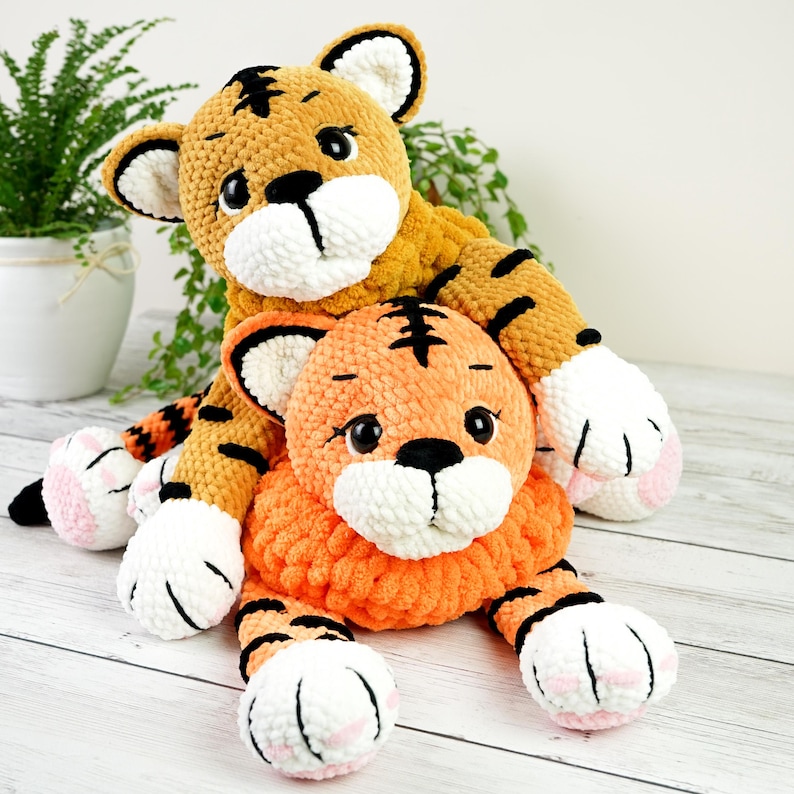 May include: Two crocheted tiger plushies, one orange and one yellow, with black stripes. The orange tiger is lying down with its paws outstretched, while the yellow tiger is sitting on top of it.
