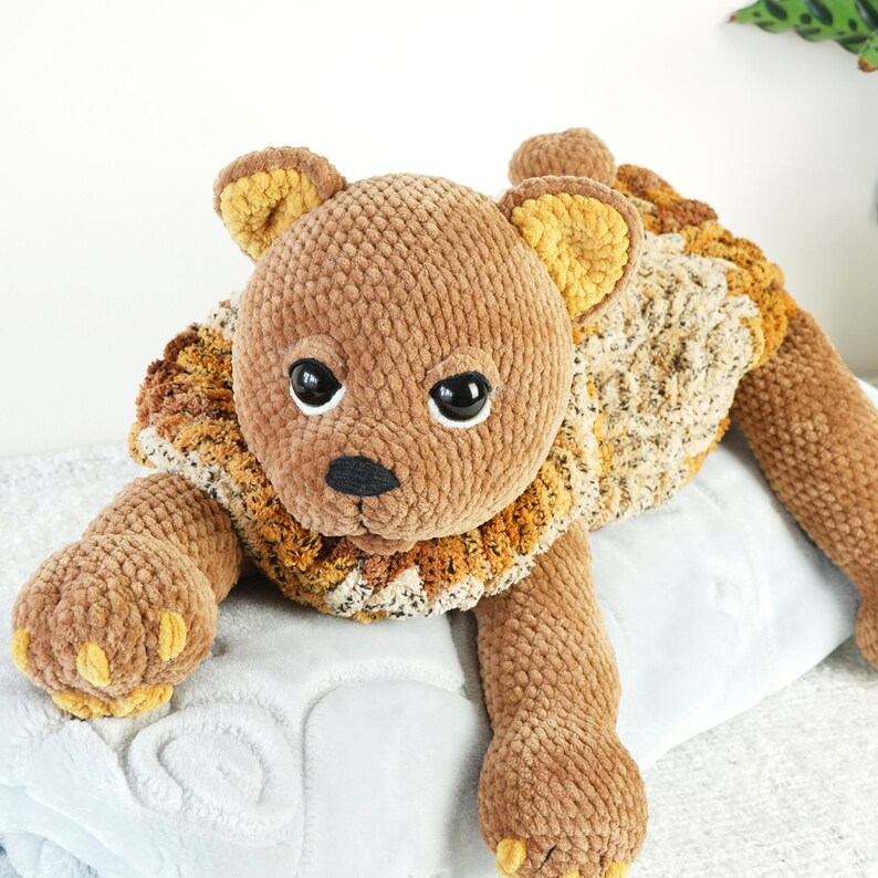 May include: A brown crocheted stuffed animal shaped like a lion laying on a white blanket. The lion has a brown body with a yellow mane and black eyes.