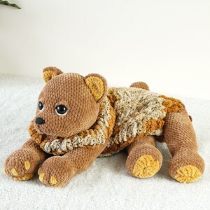 May include: A brown crocheted teddy bear with a brown and beige crocheted scarf. The bear is lying on its side with its head resting on its paws.