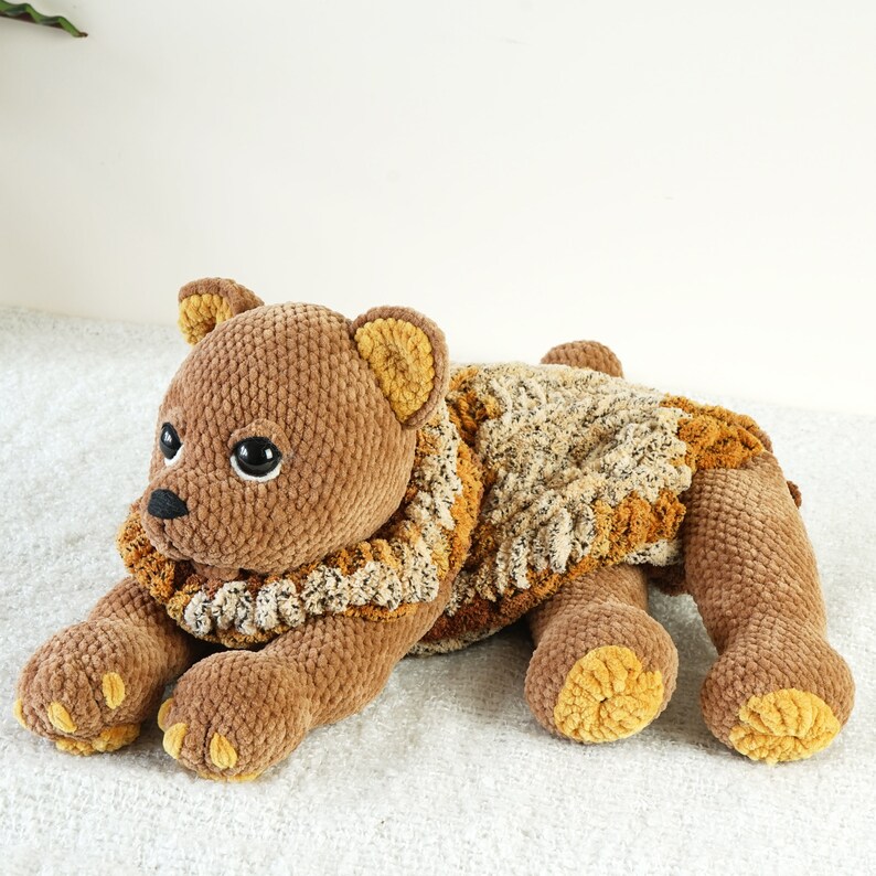 May include: A brown crocheted teddy bear with a brown and beige crocheted scarf. The bear is lying on its side with its head resting on its paws.