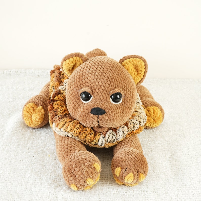 May include: A brown crocheted lion plush toy with a yellow and brown mane. The lion has black eyes and a black nose.
