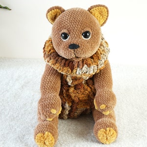 May include: A brown crocheted teddy bear with a brown and beige crocheted collar. The bear has black eyes and a black nose.