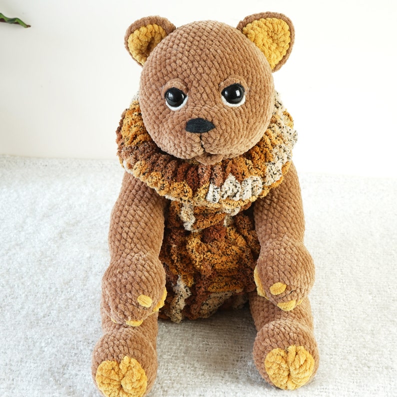 May include: A brown crocheted teddy bear with a brown and beige crocheted collar. The bear has black eyes and a black nose.