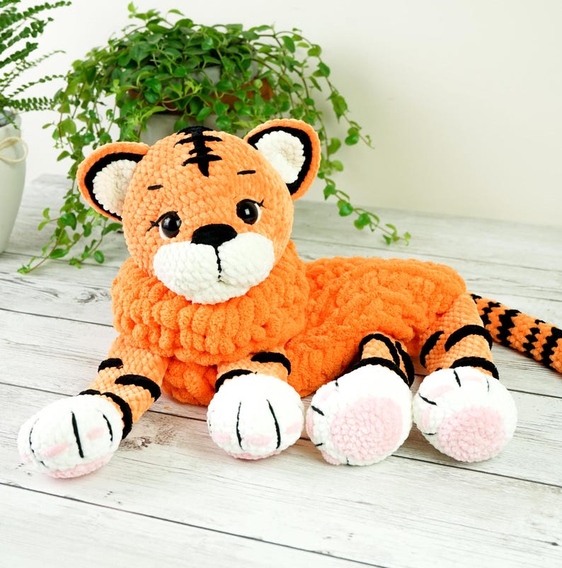 May include: A crocheted orange and black tiger plush toy with white and pink paw pads. The toy is lying on a light wood surface.