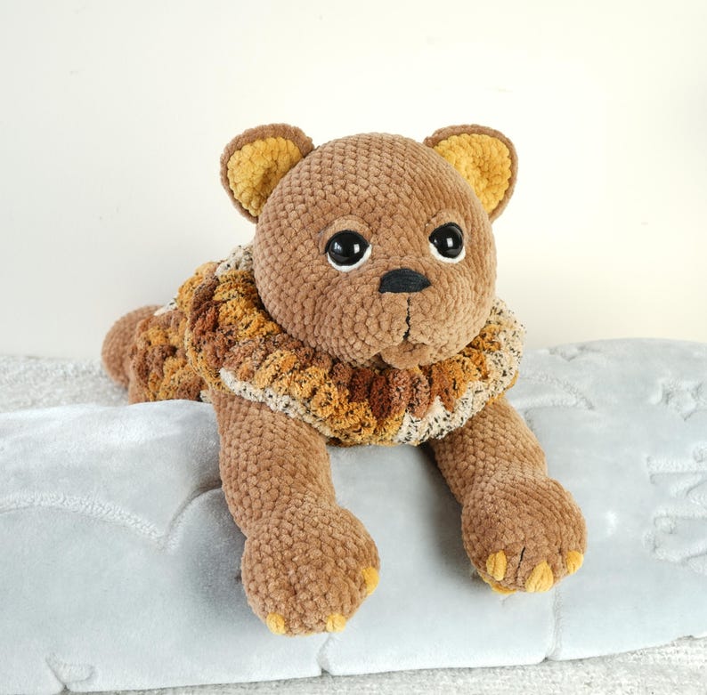 May include: A brown crocheted lion plush toy with a brown and yellow mane. The lion is lying on a gray blanket.
