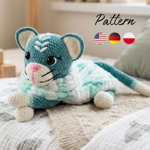 Könnte beinhalten: Ein gehäkeltes Plüschtier in Form eines Tigers aus blaugrünem, weißem und hellgrünem Garn. Das Spielzeug hat schwarze Sicherheitsaugen, eine rosa gestickte Nase und weiße Innenohren. Das Wort Pattern steht in schwarzer Schrift über drei Flaggensymbolen.