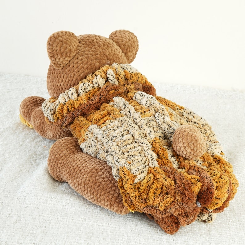 May include: A brown crocheted teddy bear wearing a brown, beige, and cream crocheted sweater. The bear is lying on its back with its legs in the air.