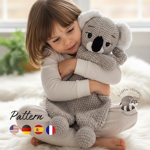 May include: A gray crocheted koala plush toy with a black nose and white fuzzy ears. The toy features a textured, knotted stitch pattern throughout its body. The text "Little Sleepy Raccoon" is printed near the bottom right with a raccoon graphic.