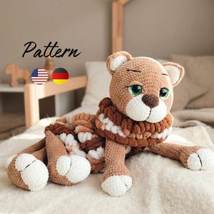 Könnte beinhalten: Ein gehäkeltes Plüschtier in Form eines Löwen mit hellbraunem Fell, einer weißen Schnauze und weißen Pfotenspitzen. Das Spielzeug hat große grüne Augen, eine dunkelbraune Nase und eine dicke, strukturierte Mähne aus braunen und weißen Garnschlingen.