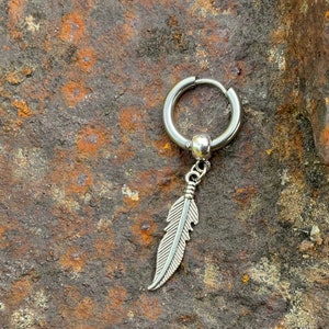 Peut inclure: Boucle d'oreille argentée avec un pendentif en forme de plume. La boucle d'oreille est dotée d'une fermeture à charnière et d'une petite perle ronde. Le charme de la plume est détaillé avec des lignes gravées. Fabriqué en métal brillant.