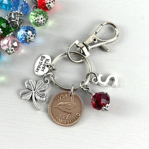 May include: A silver-toned keychain with a lobster clasp, a key ring, and various charms. Charms include a four-leaf clover, a coin with a bird, a red faceted bead, a letter "S", and a charm that says "Happy Birthday". Colorful faceted beads are visible in the background.