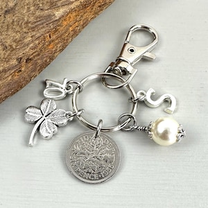 May include: Silver-toned keychain with a clover charm, the number 70, and the letter "S". It also features a vintage coin and a pearl-like bead. The keychain is attached to a silver clasp.