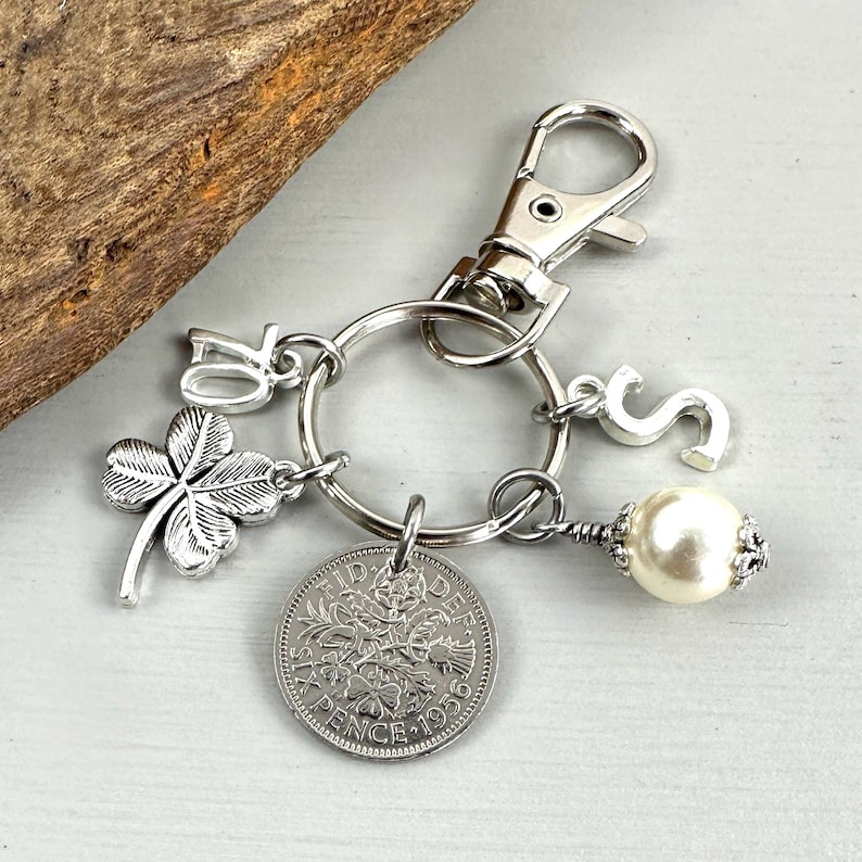 May include: Silver-toned keychain with a clover charm, the number 70, and the letter "S". It also features a vintage coin and a pearl-like bead. The keychain is attached to a silver clasp.